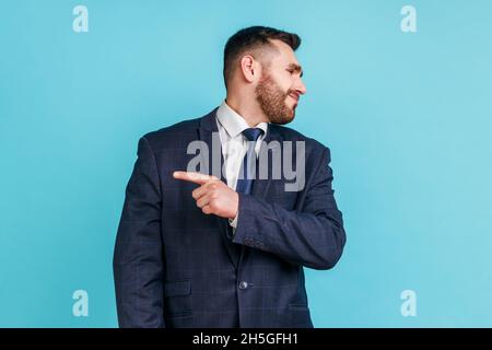 Sortez.Portrait d'un homme en colère avec une barbe portant un costume sombre regardant de côté et pointant le doigt d'une autre manière, demandant à partir, expression insatisfaite.Studio d'intérieur isolé sur fond bleu. Banque D'Images