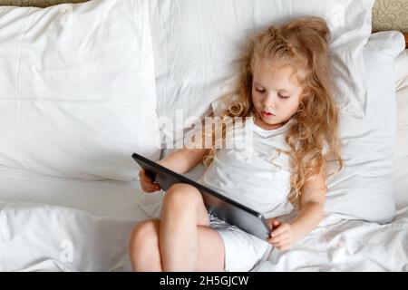 Serious attentif Caucasian Girl utilisez une tablette pour regarder des vidéos et des dessins animés.Magnifique enfant fille Portrait utilisant un ordinateur tablette couché dans le lit.Petit Banque D'Images