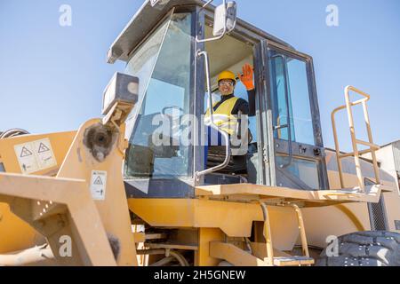 Homme joyeux assis dans le tracteur et faisant le geste bonjour Banque D'Images