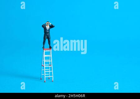 Homme d'affaires utilisant des jumelles se tenant au-dessus de l'échelle pliante. Photo miniature de jouets de personnes minuscules. Isolé sur fond bleu. Banque D'Images