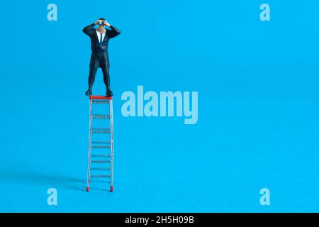 Homme d'affaires utilisant des jumelles se tenant au-dessus de l'échelle pliante. Photo miniature de jouets de personnes minuscules. Isolé sur fond bleu. Banque D'Images