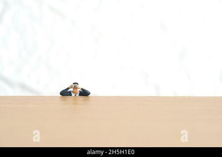 Homme d'affaires utilisant des jumelles se cachant sur un bureau en bois.Photo miniature de jouets de personnes minuscules. Isolé sur fond blanc. Banque D'Images