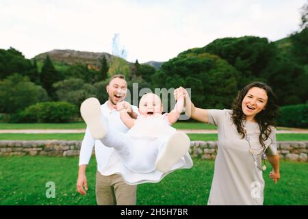Budva, Monténégro - 17.04.2017: Maman et papa souriant balancent une fille rieuse par les bras dans un jardin vert.Villa Milocer, Monténégro.Vue avant Banque D'Images
