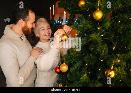 Joyeux jeune couple en amour décorant l'arbre de Noël avec des boules brillantes colorées près de la cheminée dans le salon sombre confortable. Banque D'Images