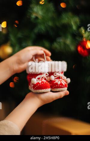 Gros plan d'une jeune femme méconnue tenant dans les mains de petites bottes rouges de noël sur fond de décoration d'arbre de Noël et de lumières de bokeh de célébration. Banque D'Images