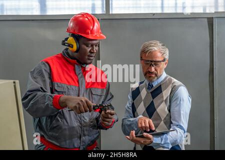 Les cotravailleurs industriels multiraciaux se tenant debout et parlant du processus de production.Black Worker Measuring avec pied à coulisse. Banque D'Images