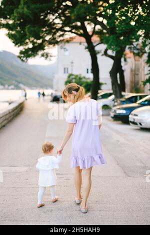 Maman conduit sa petite fille par la main le long du remblai de la ville de Perast, à côté de voitures et d'arbres.Vue arrière Banque D'Images