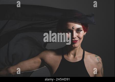 REDHEAD femme avec un voile noir sur sa tête dans un studio sombre. Une jeune fille avec un contour d'oreille dans son nez sourit sly. Maquillage de sorcière. Costume d'Halloween. Banque D'Images