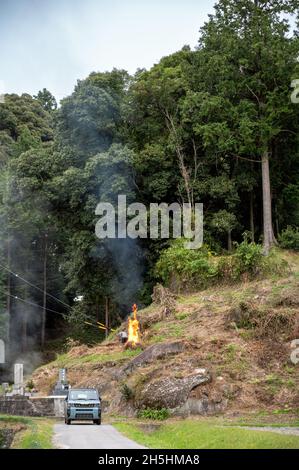 Agriculteur japonais brûlant de l'herbe sèche sur la pente de la montagne.La fumée du feu passe dans l'air.Tombes et une voiture au premier plan. Banque D'Images