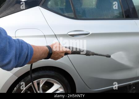 gros plan de la main de mans, tenant une buse de nettoyeur haute pression, laver à la main une voiture argentée à l'extérieur sur l'allée lors d'une journée ensoleillée Banque D'Images