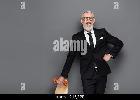 homme d'affaires heureux d'âge moyen en costume et lunettes tenant la planche tout en posant avec la main sur la hanche sur le gris Banque D'Images