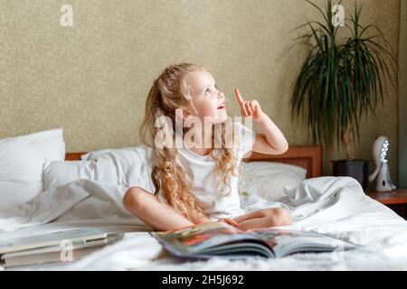 Très belle petite fille intelligente pointant vers le haut par le doigt tout en lisant le livre sur le lit le matin pendant les devoirs lire étude imaginer dans la chambre à coucher dedans Banque D'Images