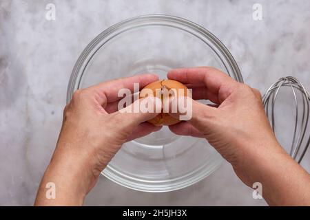 Vue de dessus de la femme craquant à la main l'oeuf brun cru dans un bol en verre sur la surface en marbre Banque D'Images