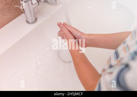 Un garçon caucasien se lave les mains dans une salle de bain légère avec du savon et de l'eau du robinet Banque D'Images