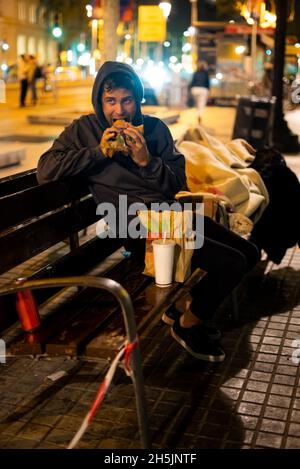 Un homme sans-abri affamé à capuchon qui regarde loin et qui mord le hamburger tout en étant assis sur le banc près d'une couverture la nuit Banque D'Images