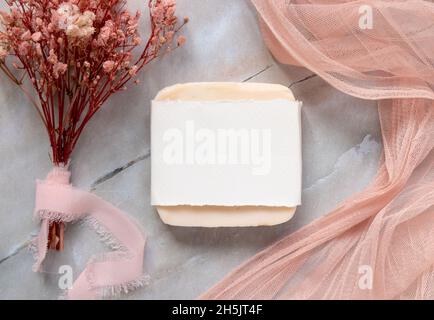 Bar à savon fait main, posé sur une table en marbre avec emballage en papier, tissus de tulle rose et fleurs séchées, vue du dessus.Maquette de savon plat la Banque D'Images