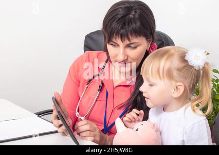 Le médecin pédiatre présente des dessins animés sur la tablette d'une petite fille de 3-4 ans.Le concept de contact psychologique avec un enfant chez un médecin Banque D'Images