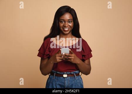 Positive jeune afro-américaine femme avec le plus récent téléphone portable regardant la caméra et souriant sur fond beige studio, en utilisant de belles rencontres, divertissement Banque D'Images