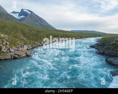 Bleu eau sauvage des rapides de la rivière Vuojatadno avec le mont Akka, massif d'Ahkka avec neige et glacier et forêt de bouleau.Sartique du nord béatiful Banque D'Images