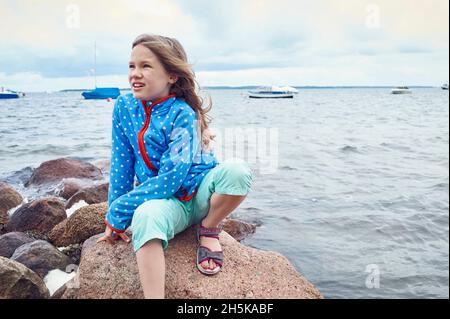 Une jeune fille de 8 ans assise sur quelques rochers dans l'eau par un jour nuageux, regardant autour, quelques petits bateaux à moteur en arrière-plan, la mer Baltique; l'Allemagne Banque D'Images