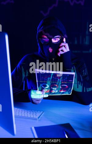 Hacker en balaclava parler sur smartphone et tenir le papier près de l'ordinateur sur fond noir Banque D'Images