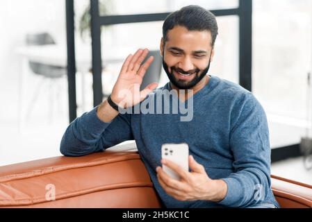 Un jeune homme indien de l'est à barbe heureux tenant un téléphone portable et en signe de bonjour, faisant un appel vidéo, un homme hispanique heureux de voir un interlocuteur en ligne, utilisant un smartphone pour la connexion virtuelle, parlant en ligne Banque D'Images