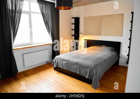 Chambre simple dans le design industriel avec mur en brique blanche, plancher en bois, plafond en béton, rideaux en coton noir et tête de lit à panneaux dans le moderne Banque D'Images