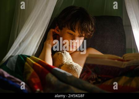 Un garçon d'entre-deux est assis sur une chaise avec un ours en peluche lisant par une lamtrette Banque D'Images