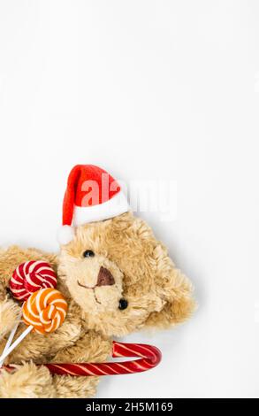 Photo verticale avec ours en peluche Teddy dans un chapeau rouge du Père Noël sur une oreille, tenant la canne à sucre et deux sucettes rayées dans ses pattes.Fond blanc Banque D'Images