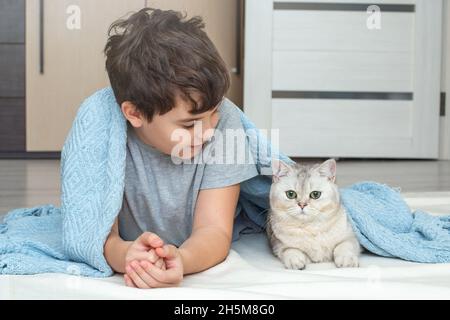 le beau garçon adolescent, brunet, se trouve avec un chat blanc sur le sol sous une couverture bleue Banque D'Images