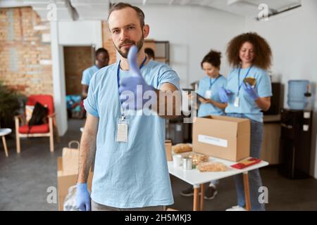 Portrait du volontaire de sexe caucasien en uniforme bleu et gants de protection montrant les pouces vers le haut, regardant l'appareil photo.Tri en équipe, emballage des articles Banque D'Images