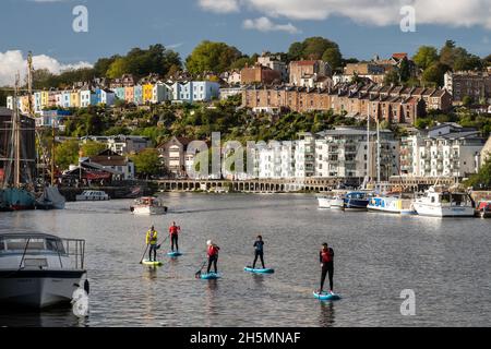 Un groupe de paddleboardeurs traversent le port flottant de Bristol, avec les maisons de Clifton Vale derrière. Banque D'Images