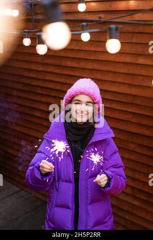Une jeune femme heureuse en vêtements d'extérieur avec des spameurs dans la rue sur le fond d'un mur en bois se réjouit des vacances du nouvel an Banque D'Images