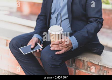 Beau et réussi entrepreneur mûr reposant avec une tasse de café en plein air profiter du temps ensoleillé du printemps et de l'utilisation du smartphone. concept de pause café Banque D'Images