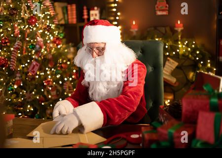 Santa Clause prépare des cadeaux pour les enfants pour Noël à son bureau à la maison tout en ouvrant des listes de souhaits Banque D'Images