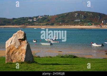 Une pierre debout solitaire sur l'herbe au-dessus du chenal bleu de la mer entre Tresco et Bryher avec de petits bateaux amarrés dans les eaux peu profondes des îles Banque D'Images