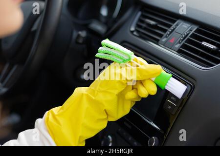 Une main dans des gants en caoutchouc jaune essuie le tableau de bord d'une voiture de la poussière avec une brosse spéciale lors d'un jour d'automne brillant.Mise au point sélective.Gros plan Banque D'Images