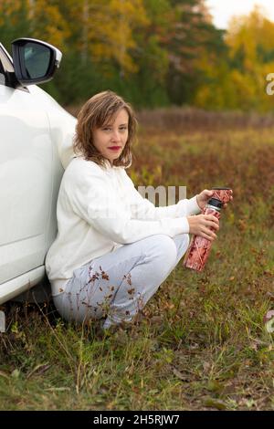 Une jolie jeune femme à capuche blanche est assise près d'une voiture blanche avec un thermos dans ses mains et boit du thé chaud le jour de l'automne sur fond Banque D'Images