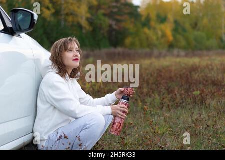 Une jolie jeune femme à capuche blanche est assise près d'une voiture blanche avec un thermos dans ses mains et boit du thé chaud le jour de l'automne sur fond Banque D'Images