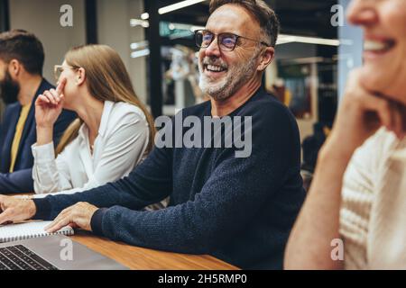 Homme d'affaires mature assistant à une réunion avec ses collègues.Bon homme d'affaires mûr riant gaiement tout en étant assis dans une salle de réunion avec son équipe.G Banque D'Images
