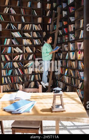 étudiant afro-américain en lunettes debout sur une échelle en bois tout en lisant le livre près de blocs-notes flous sur le bureau Banque D'Images