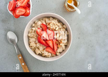 Porridge de flocons d'avoine avec tranches de fraises, amandes de noix et miel dans un bol sur table grise. Alimentation saine, diète, concept de nourriture végétarienne. Placer pour tex Banque D'Images