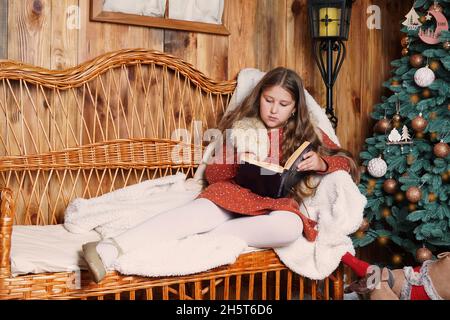 Jeune fille souriante aux cheveux longs allongé sur un canapé en osier, lisant un livre, tenant une tasse de boisson chaude près de l'arbre de Noël décoré.Fille à Noël. Banque D'Images