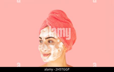 Jeune femme heureuse avec une serviette rose sur sa tête lave le visage, nettoie la peau avec de la mousse de savon.Concept de routine du matin et de soin de la peau Banque D'Images