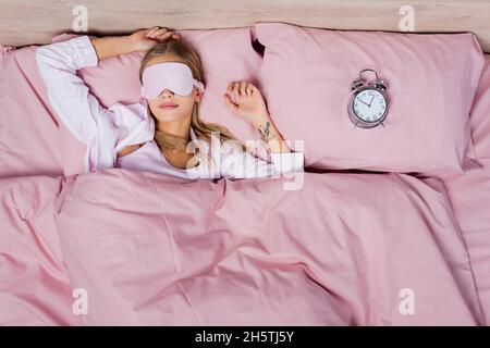 Vue de dessus de la femme dans le masque de sommeil et le pyjama couché près du réveil sur le lit Banque D'Images