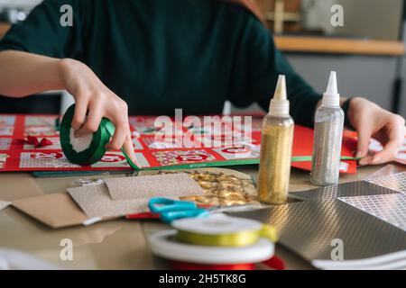 Gros plan des mains d'une jeune femme méconnue utilisant un ruban de Noël pour encadrer le tableau avec des enveloppes de calendrier de l'Avent à la maison. Banque D'Images