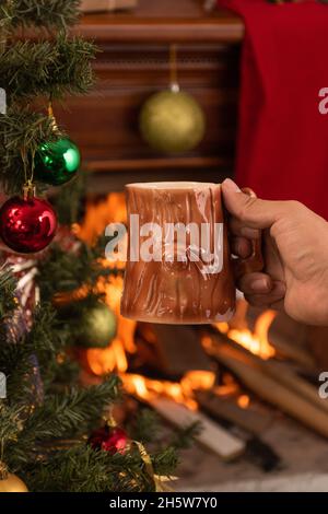 main tenant une tasse originale avec un design en bois, avec une boisson chaude, en arrière-plan une cheminée en bois avec une flamme, appréciant à la maison en hiver Banque D'Images