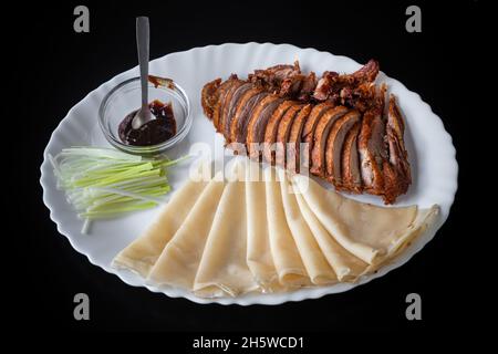 Canard de Pékin, canard croustillant frit à la chinoise, servi avec une sauce hoisine, des crêpes et du concombre, dans une assiette blanche sur fond noir. Banque D'Images
