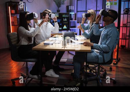 Groupe de quatre collègues multiraciaux portant un casque VR lors d'une réunion de travail au bureau.Partenaires d'affaires assis au bureau et touchant l'écran virtuel. Banque D'Images