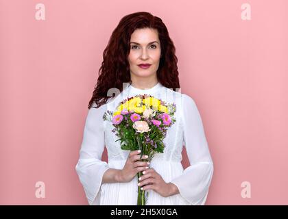 Femme tenant des fleurs devant sa poitrine avec fond rose.Portrait de studio romantique de belle femme en robe blanche tenant le bouquet de fleurs. Banque D'Images